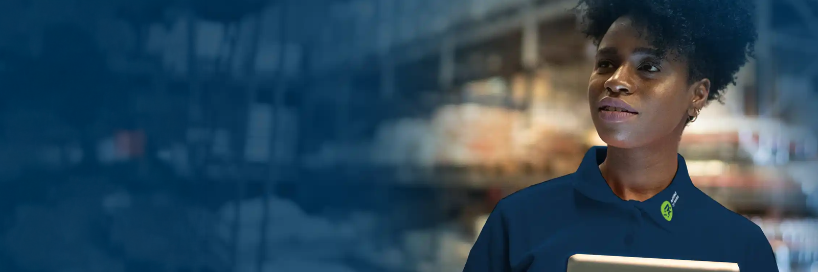 mitarbeiterin in dunkelblauem poloshirt haelt ein tablet in einem lager mit hohen regalen; linker bildbereich ist in blau getoent; das motiv steht fuer einen modernen fulfillment-dienstleister der logistik digital und transparent steuert