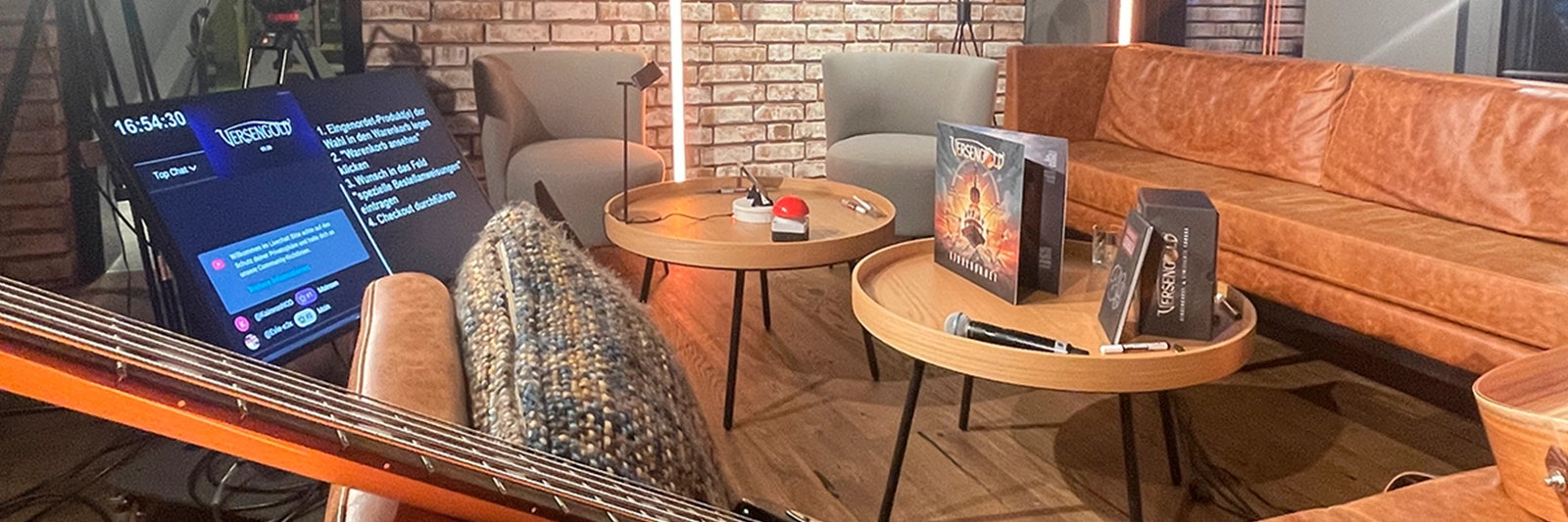 youtube-live-set in lounge-optik mit sofas, runden tischen und teleprompter-monitor; album-artwork und instrumente liegen bereit fuer das versengold live-event