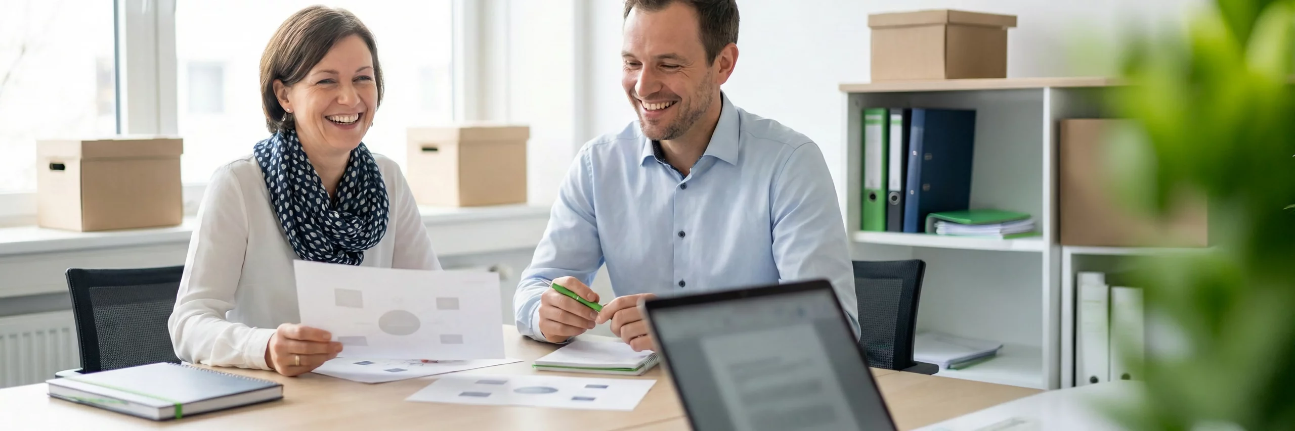 zwei personen mit europaeischem erscheinungsbild sitzen an einem tisch und betrachten diagramm-ausdrucke ohne text; beide laecheln konzentriert, das motiv steht fuer transparentes reporting und nachhaltigkeits-kpis im fulfillment